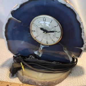 May include: A decorative clock with a round face and gold-colored trim set into a polished blue and white agate stone. The clock has black hands and a black power cord. The base is a matching agate stone.