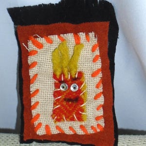 May include: A handmade felt art piece featuring a whimsical creature with yellow tufts and googly eyes. The creature is stitched onto a beige fabric, framed by orange and black felt, and accented with orange embroidery.