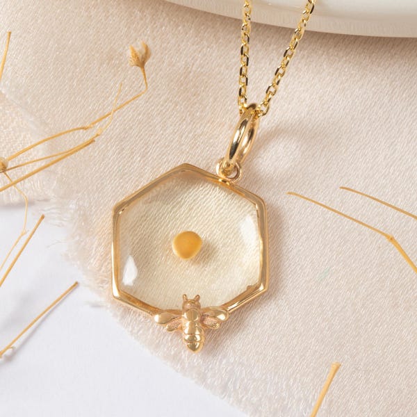 Puede incluir: Un collar dorado con un colgante hexagonal. El colgante tiene un centro transparente con un pequeño objeto redondo y amarillo en su interior. Una pequeña abeja está en la parte inferior del colgante. El collar está sobre una superficie blanca.