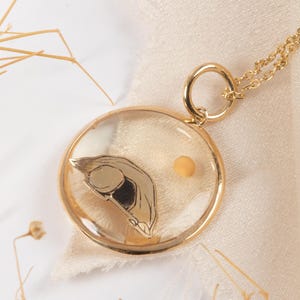 May include: A gold-colored pendant necklace with a clear, circular resin-filled charm. Inside the charm is a gold-colored, stylized seed design and a small, round, yellow object. The necklace chain is also gold-colored.