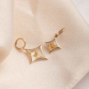Könnte beinhalten: Zwei goldene sternförmige Anhänger mit klaren Glaseinsätzen, die kleine, runde, gelbe Objekte enthalten. Die Anhänger hängen an einem hellbeigen Stoffhintergrund.