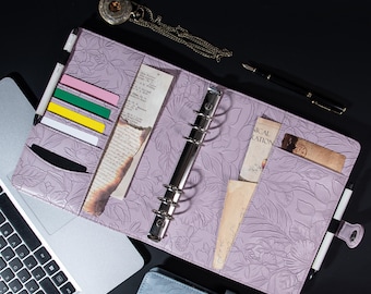 Op de afbeelding: Een lavendelkleurige leren planner met een reliëf van bloemen. De planner heeft een ringband-systeem en meerdere vakken om kaarten, notities en andere items in te bewaren. De planner ligt open op een laptop.