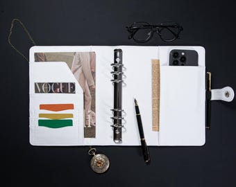 Puede incluir: Planificador encuadernado en cuero blanco con un archivador de anillas, un bolígrafo, un teléfono y un bolsillo con la palabra "VOGUE" impresa.