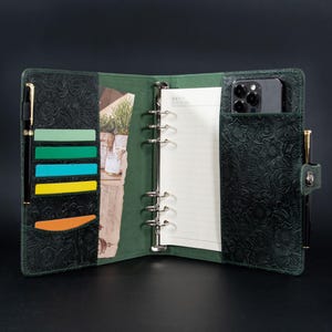 May include: Open green leather binder with floral embossing. The binder has multiple card slots, a notepad, and a pocket holding a smartphone. A pen is secured on each side. The binder is open, revealing the interior.