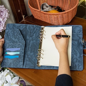 May include: A blue leather-like planner cover with an embossed floral pattern, featuring a six-ring metal binder mechanism, multiple interior card slots, and a grid-lined paper insert. A black and gold fountain pen rests on the open page.