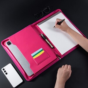 May include: A pink leather zippered portfolio with a notepad, pen, and card slots. The portfolio is open and a hand is writing on the notepad.