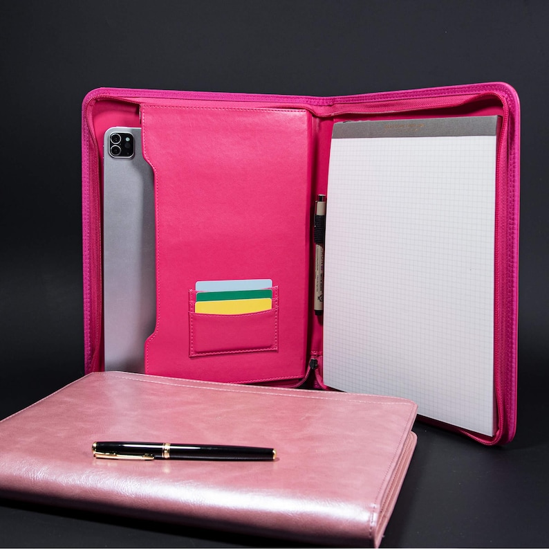 May include: A pink leather zippered folder with a notepad, pen, and card slots. The folder is open and the notepad is visible. The folder is on a black surface.