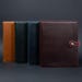 May include: Four rectangular leather notebook covers arranged in a row. The covers are tan, dark green, navy blue, and deep burgundy. Each cover features a smooth matte finish and stitched edges. The burgundy cover has a tab closure with a metal snap.