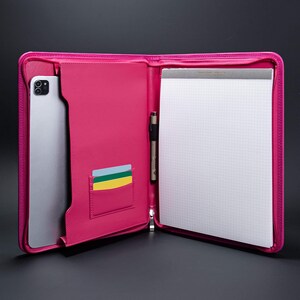 May include: A pink leather zippered portfolio with a tablet, pen, and card slots. The portfolio is open and the interior is lined with a white gridded notepad.