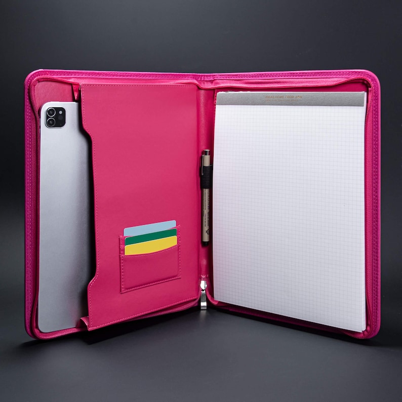 May include: A pink leather zippered portfolio with a tablet, pen, and card slots. The portfolio is open and the interior is lined with a white gridded notepad.
