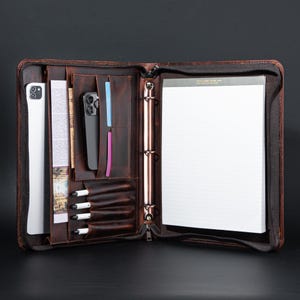 May include: A brown leather zippered binder with a built-in notepad, pen holders, and a pocket for a phone or tablet. The binder has a ring mechanism for holding papers.