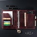 May include: A dark red leather A5 binder featuring a six-ring metal mechanism, multiple interior pockets holding cards and papers, a smartphone slot, and a pen loop. A silver-toned pocket watch hangs from the bottom, and two pens rest above.