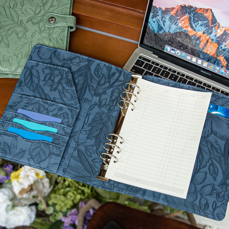 May include: A blue floral embossed leather binder featuring a six-ring metal mechanism, internal card slots holding three colored cards, and a white lined paper insert. The binder is open, revealing its textured surface and interior pockets.