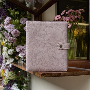 May include: A light pink rectangular binder featuring an embossed floral pattern across the entire front cover. The binder includes a matching pink strap closure secured with a single metallic button fastener centered on the right side edge.