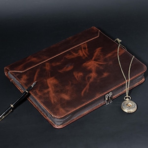 May include: A brown leather portfolio with a zip closure, a black pen, and a vintage pocket watch with a gold chain. The portfolio has a distressed finish and a flap closure. The pocket watch has an ornate design.