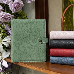 May include: A sage green leather planner with an embossed floral pattern and a snap closure, displayed alongside a stack of four additional planners in light pink, black, red, and blue, each featuring the same embossed floral design.