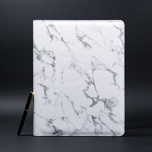 Puede incluir: Un cuaderno de cuero blanco con un patrón de mármol. El cuaderno está cerrado y acostado sobre una superficie negra.