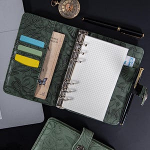 Puede incluir: Agenda de cuero verde abierto con relieve floral, un bloc de notas cuadriculado y una carpeta plateada. El planificador tiene ranuras para tarjetas con tarjetas de colores, un marcador con una mariposa y un bolígrafo negro. Un reloj de bolsillo y un portátil están en el fondo.