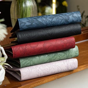 May include: A stack of five rectangular journals with embossed floral patterns on the covers. The journals are colored in blue, black, red, sage green, and light lavender. Each journal features a consistent botanical design pressed into the material.