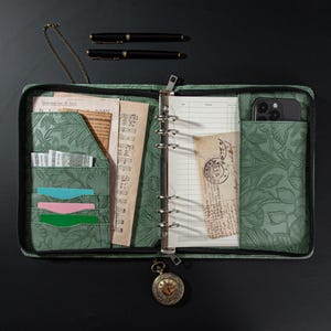 Op de afbeelding: Open groene leren organizer met een bloemreliëf, met papieren, kaarten en een smartphone. Twee zwarte pennen liggen erboven. Een vintage zakhorloge hangt aan de rits.