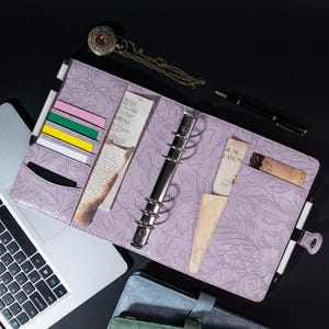 Puede incluir: Una agenda de cuero de color lavanda con un diseño estampado de flores. La agenda tiene un sistema de encuadernación con anillas y varios bolsillos para guardar tarjetas, notas y otros artículos. La agenda está abierta y sobre un ordenador portátil.