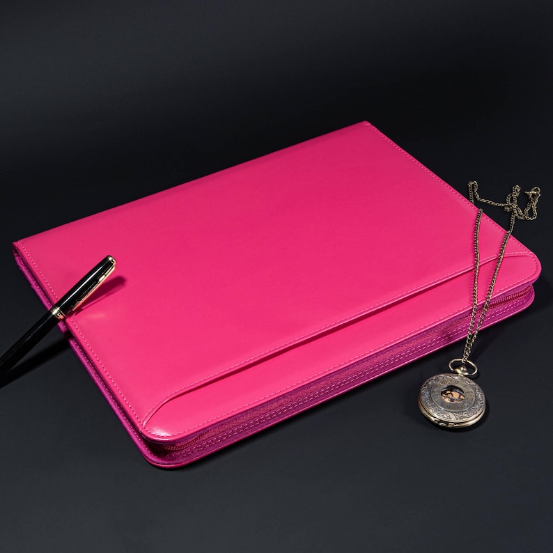 May include: A bright pink leather zippered portfolio with a gold chain and pocket watch attached. A black pen is lying on top of the portfolio.