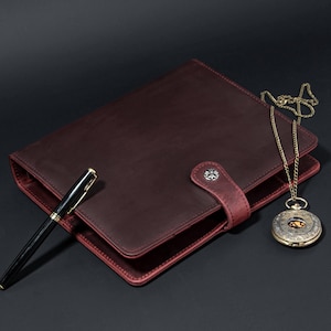 May include: A burgundy leather journal with a pen and a vintage pocket watch on a chain. The journal has a strap closure with a decorative button. The pen is black with gold accents.