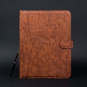 May include: A brown leather folder with a floral embossed design. The folder has a flap closure and a pen holder.