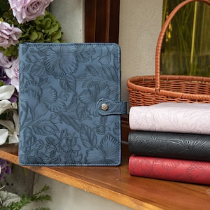 May include: A blue rectangular planner with an embossed floral pattern and a single snap button closure. Beside it, a stack of three similar planners in light pink, black, and red, all featuring the same embossed floral design and texture.