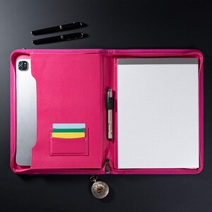 May include: A pink leather portfolio with a zipper closure, a pen holder, and a card slot. The portfolio is open to reveal a blank, gridded notepad.