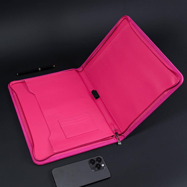 May include: A pink zippered portfolio case with a black zipper pull. The case is open and has a pocket on the inside. A black pen and a black smartphone are on the table.