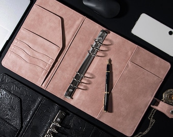 Op de afbeelding: Twee open lederen planners met ringbinders. De ene planner is zwart met een reliëfmotief van bloemen en de andere is roze. Een pen en een computermuis liggen op de tafel.