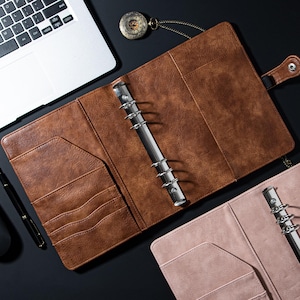 May include: Two leather-bound ring binders, one brown and one pink, with pockets and a metal ring mechanism. Both binders are open and lying flat on a black surface.
