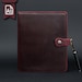 May include: A dark burgundy A5 leather notebook cover with visible stitching along the edges. The cover features a side strap closure secured by a silver-toned metal button with a floral pattern. A black ballpoint pen is tucked into the side.