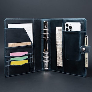 May include: A dark blue leather bound planner with a ring binder system. The planner has a pocket for a phone, a pen holder, and several card slots. The planner is open to reveal a page with a newspaper clipping.