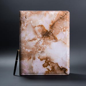 Puede incluir: Una cubierta de cuaderno de cuero de color rosa y marrón con un diseño de mármol y un bolígrafo dorado.