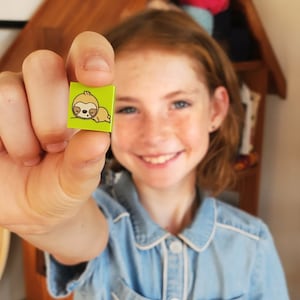 May include: A small, square, lime green tile featuring a cartoon sloth illustration. The sloth is light brown with a white face and is lying down. The tile is held up by a person.