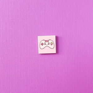 May include: A light pink square sticker featuring a cartoon video game controller with a smiling face. The controller has red and yellow buttons and a red directional pad. The background is a textured purple surface.