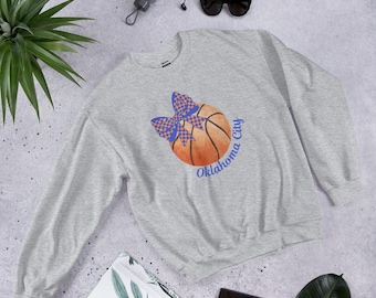 Może przedstawiać: Jasnoszara bluza z nadrukiem koszykówki i niebiesko-pomarańczową kokardą. Na koszykówce widnieje napis "Oklahoma City" w niebieskim kolorze. Bluza wykonana jest z miękkiego materiału.