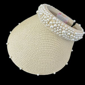 May include: A beige straw sun visor with a wide brim and a band of white pearls around the crown.