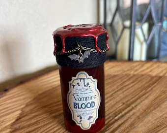 May include: A small glass bottle filled with red liquid, labeled "Vampire Blood." The bottle is topped with a black band and a red wax seal. A small bat charm hangs from a chain around the neck of the bottle. The bottle sits on a wooden surface.
