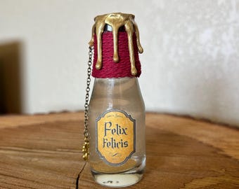 May include: A small glass bottle with a gold wax seal and a red string tied around the neck. The bottle is filled with a clear liquid and has a label that reads "Felix Felicis".