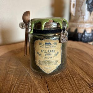 Puede incluir: Un frasco de vidrio lleno de purpurina verde y etiquetado "Polvo de Floo" con instrucciones de uso. El frasco tiene un sello de cera verde y una cuchara de madera.