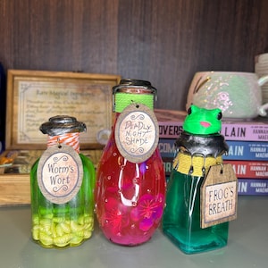 May include: Three glass bottles with decorative labels. The left bottle is green with yellow worm-like shapes and a label reading Worm's Wort. The center bottle is red with pink flowers and a label reading Deadly Night Shade. The right bottle is green with a frog topper and a label reading Frog's Breath.
