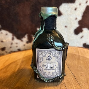 Puede incluir: Una pequeña botella de vidrio con una etiqueta negra que dice "Basilisk Venom" y un sello de cera verde. La botella está envuelta en una serpiente verde y está sobre una superficie de madera.