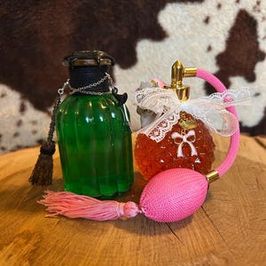 Könnte beinhalten: Zwei antike Glasparfümflakons mit dekorativen Stopfen und Quasten. Die Flasche links ist grün und hat einen schwarzen Stopfen mit einer Kette und einem kleinen Besen-Anhänger. Die Flasche rechts ist orange und hat eine rosa Quaste und eine weiße Schleife aus Spitze.