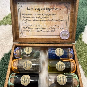 Könnte beinhalten: Eine Holzkiste mit einem Etikett mit der Aufschrift "Seltene Magische Zutaten" und sechs kleinen Glasbehältern, die mit verschiedenen Zutaten gefüllt sind, darunter Mondstein, Dillany-Blätter, Blutegel-Saft, Mischlingsfell, Troll-Schleim und Spinnen-Gift.