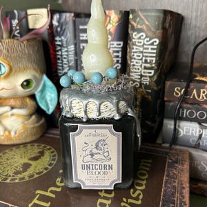 Op de afbeelding: Een vierkante glazen pot gevuld met donkere vloeistof, voorzien van een etiket met de tekst Moonlight Apothecary Unicorn Blood Pure Extract. Het deksel is versierd met zilveren druppels, een witte gedraaide kaars, blauwe kralen en grijs mos.