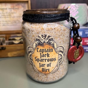 Op de afbeelding: Een glazen pot gevuld met lichtgekleurd zand, afgesloten met een donkerhouten deksel en zwart touw. Een etiket vermeldt Captain Jack Sparrows Jar of Dirt. Een zwart koord met twee houten kralen en een rood waszegel hangt aan de rand van de pot.