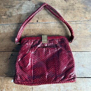 May include: A vintage red handbag with a snakeskin pattern. The bag has a structured shape, a top handle, and a gold-colored clasp. The bag is made of a textured material and sits on a wooden surface.